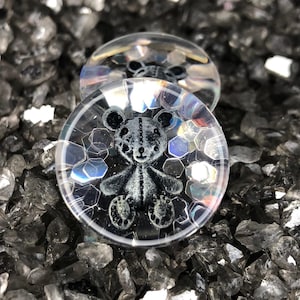 May include: Two clear glass cabochons with a black and white teddy bear design. The cabochons have a holographic finish with a hexagonal pattern.