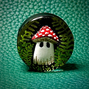 Könnte beinhalten: Ein kleiner, weißer Pilz mit roten und weißen Punkten auf dem Hut, umgeben von grünen Farnen. Der Pilz ist auf einem runden, schwarzen Hintergrund gemalt.