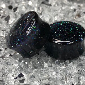 May include: Two black ear gauges with iridescent glitter embedded in the resin. The gauges are round and have a smooth, polished finish.