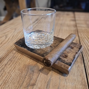 Puede incluir: Un posavasos de madera con un vaso y un cigarro apoyados en él. El posavasos está hecho de madera reciclada y tiene un acabado marrón oscuro.