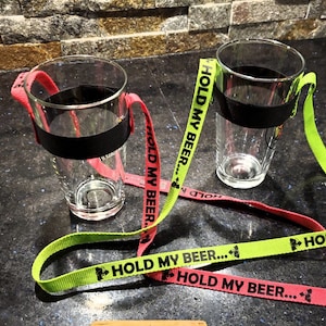 K&ouml;nnte beinhalten: Zwei klare Glasbierkr&uuml;ge mit schwarzen B&auml;ndern um den oberen Rand. Jeder Krug hat ein neon-gr&uuml;nes und ein neon-rosa Band daran befestigt. Die B&auml;nder haben den Text "HOLD MY BEER..." aufgedruckt.
