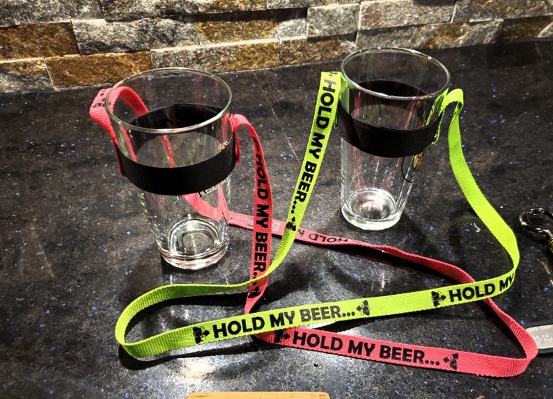 K&ouml;nnte beinhalten: Zwei klare Glasbierkr&uuml;ge mit schwarzen B&auml;ndern um den oberen Rand. Jeder Krug hat ein neon-gr&uuml;nes und ein neon-rosa Band daran befestigt. Die B&auml;nder haben den Text "HOLD MY BEER..." aufgedruckt.