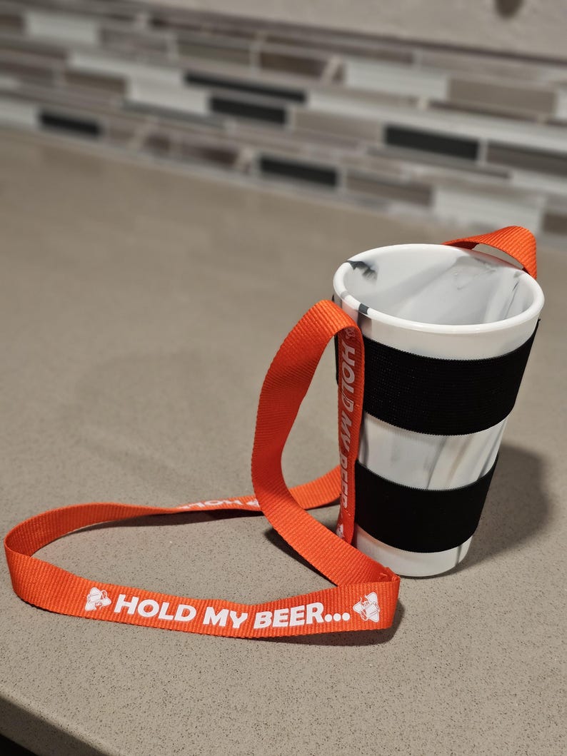 K&ouml;nnte beinhalten: Orangefarbener und wei&szlig;er "Hold My Beer..."-Gurt mit einem schwarz-wei&szlig; gestreiften Becherhalter.