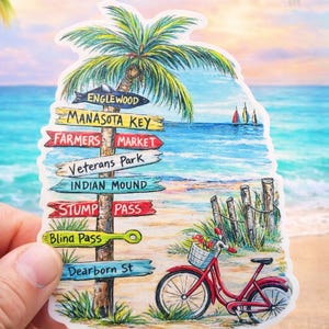 May include: A colorful sticker featuring a beach scene with a palm tree, ocean, and a red bicycle with a basket of flowers. Directional signs point to Englewood, Manasota Key, Farmers Market, and other locations. The background shows a sunset.