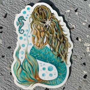Peut inclure: Un autocollant repr&eacute;sentant une sir&egrave;ne avec de longs cheveux bruns et turquoise, une queue turquoise et un collier de perles. Un hippocampe turquoise et des bulles sont &eacute;galement inclus dans le dessin. La sir&egrave;ne est de dos.