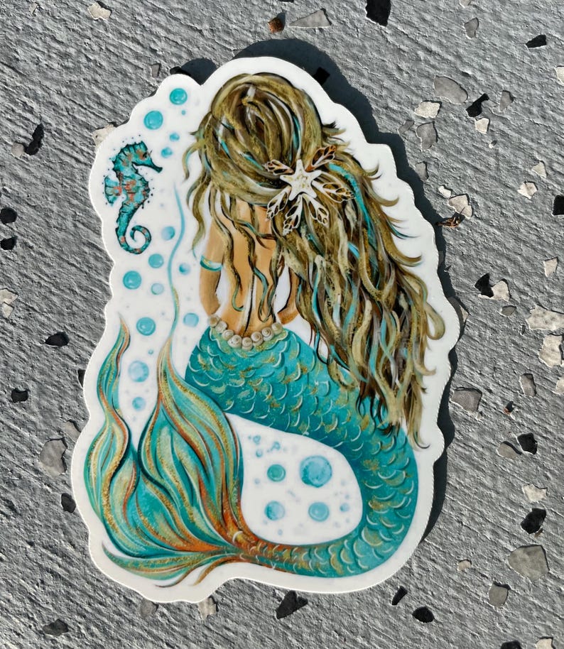 Peut inclure: Un autocollant repr&eacute;sentant une sir&egrave;ne avec de longs cheveux bruns et turquoise, une queue turquoise et un collier de perles. Un hippocampe turquoise et des bulles sont &eacute;galement inclus dans le dessin. La sir&egrave;ne est de dos.
