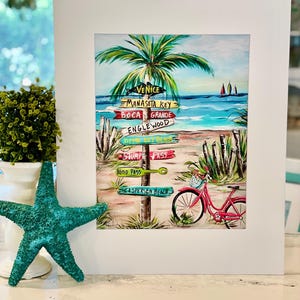Puede incluir: Una lámina de arte montada que muestra una escena de playa con una palmera, una bicicleta roja y un poste señalizador con los nombres Venice, Manasota Key, Boca Grande, Englewood, Siesta Key Beach, Stump Pass, Blind Pass y Caspersen Beach.