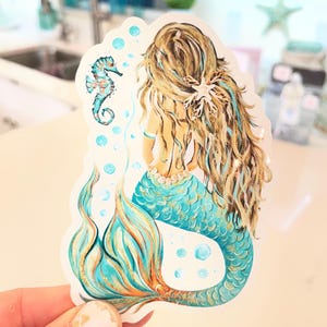 Peut inclure: Autocollant de sir&egrave;ne avec une queue turquoise et dor&eacute;e, de longs cheveux blonds et un hippocampe. La sir&egrave;ne est vue de dos, avec un collier de perles et un accessoire de cheveux en forme d'&eacute;toile de mer. Des bulles entourent la sir&egrave;ne et l'hippocampe.