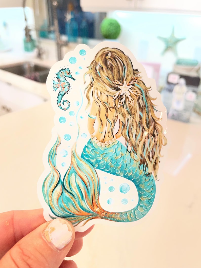 Peut inclure: Autocollant de sir&egrave;ne avec une queue turquoise et dor&eacute;e, de longs cheveux blonds et un hippocampe. La sir&egrave;ne est vue de dos, avec un collier de perles et un accessoire de cheveux en forme d'&eacute;toile de mer. Des bulles entourent la sir&egrave;ne et l'hippocampe.