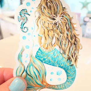 Peut inclure: Autocollant de sir&egrave;ne avec de longs cheveux blonds, une queue turquoise et un collier de perles. Un hippocampe et des bulles sont &eacute;galement repr&eacute;sent&eacute;s. L'autocollant est tenu par une main avec du vernis &agrave; ongles bleu clair.