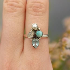 May include: A silver ring with a cluster of gemstones. The ring features a central pearl, a round turquoise stone, a pear-shaped blue stone, and an oval, translucent stone. The gemstones are set on a silver band.