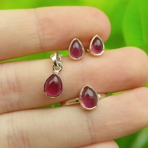 May include: A jewellery set comprising a pendant, stud earrings, and a ring. Each item features a teardrop-shaped, deep pink gemstone set in silver. The ring and earrings are small, while the pendant hangs from a silver chain.