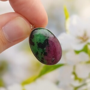 May include: An oval-shaped pendant featuring a polished zoisite stone with distinct patches of vibrant green, deep magenta, and black inclusions, set within a simple silver-toned metal frame with a small bail at the top for attachment.