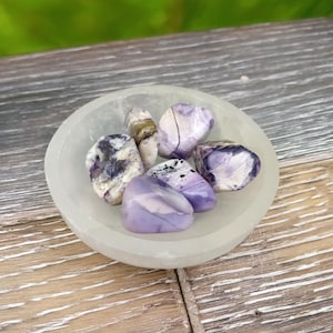 May include: A small bowl filled with tumbled amethyst stones, featuring shades of purple and white. The bowl is made of white selenite and has a smooth, polished finish.