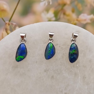 May include: Three silver pendant necklaces with oval-shaped, iridescent blue and green opal stones. The opals have a dark blue base colour with flashes of green and other colours. The pendants are displayed on a white surface.