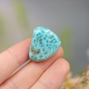May include: A polished, turquoise gemstone with a distinctive pattern of lighter blue veins. The stone has a smooth, rounded shape and a few small brown inclusions. The gemstone is held in a hand, with a blurred background.