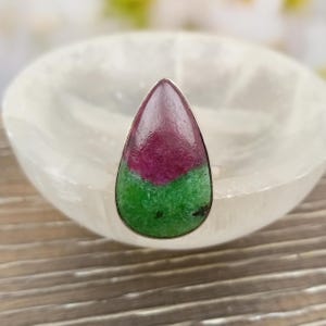 May include: A teardrop-shaped ring featuring a polished gemstone with a distinct colour split. The upper portion is a deep magenta-red, while the lower section is a vibrant, mottled green. The stone is set in a simple, thin silver-toned metal bezel.