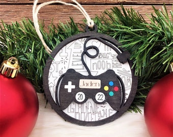May include: A round, wooden Christmas ornament featuring a black game controller design. The ornament has the name "Jaden" and the year "2022" on it. The background is white with game-related doodles. A twine loop is attached for hanging.