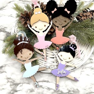 May include: Four wooden ballerina ornaments in pink, peach, light blue, and lavender tutus. Each ornament has a different hairstyle and a ribbon for hanging. One ornament is personalized with the name "Sofia" and the year "2023". The ornaments are displayed in a white basket with pine needles.