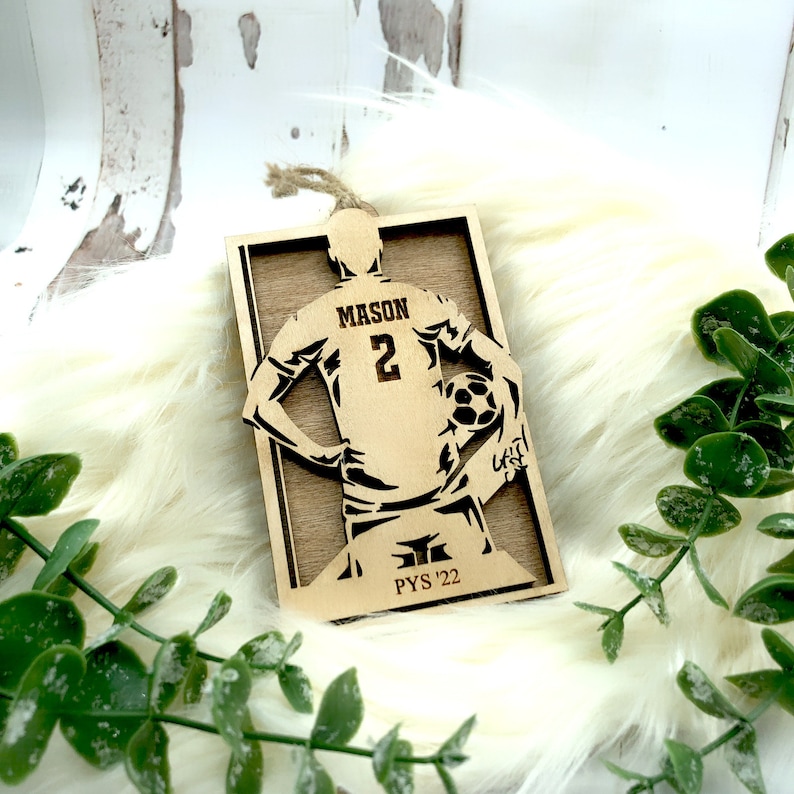 May include: Wooden soccer ornament featuring a player with the name "MASON" and the number "2" on the jersey. The ornament includes the text "PYS '22" and a soccer ball. It has a natural wood finish and a twine loop for hanging.