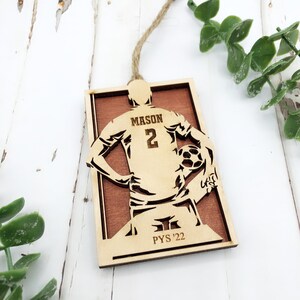 May include: A wooden ornament featuring a soccer player silhouette holding a soccer ball. The player's jersey has the name "MASON" and the number "2". The ornament is rectangular with a brown background and the text "PYS '22". A twine loop is attached for hanging.