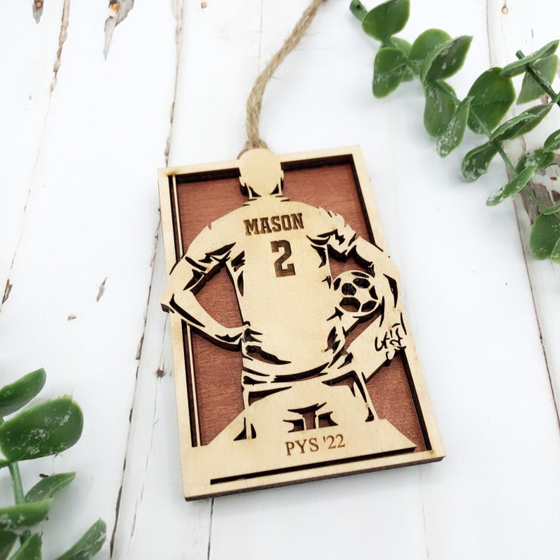 May include: A wooden ornament featuring a soccer player silhouette holding a soccer ball. The player's jersey has the name "MASON" and the number "2". The ornament is rectangular with a brown background and the text "PYS '22". A twine loop is attached for hanging.