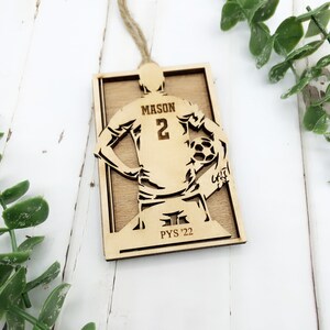 May include: A wooden soccer-themed ornament featuring a player with the name "MASON" and the number "2" on the jersey. The ornament includes the text "PYS '22" and is designed with a soccer ball. It has a natural wood finish and a twine hanger.