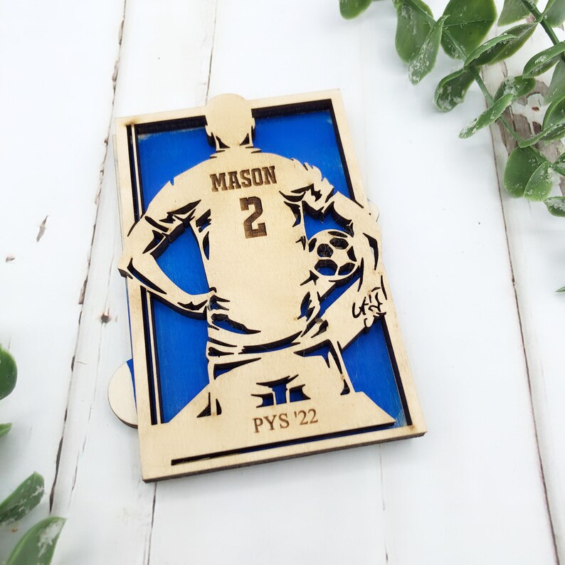 May include: A wooden soccer player figure with the name "MASON" and the number "2" on the back of the jersey. The figure is set against a blue background and includes the text "PYS '22". The soccer player is holding a soccer ball.