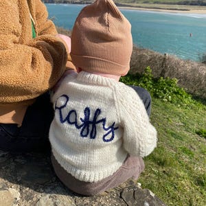 Può includere: Un bambino indossa un maglione lavorato a maglia color crema con il nome "Raffy" ricamato in blu. Il bambino indossa anche un berretto color beige e pantaloni marroni. Lo sfondo mostra un paesaggio costiero con acqua blu e colline verdi.