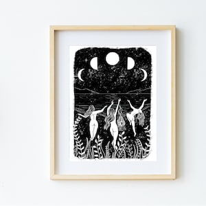 A4 art print on a white wall. The print is of three ladies dancing naked around a fire under the moon phases on the beach, surrounded by plants and leaves. Black & white print in lino style art.