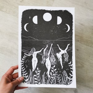 May include: Black and white linocut print of three women dancing in front of a bonfire under a night sky with three moons. The women are nude and have long hair. The print is framed by a border of plants.
