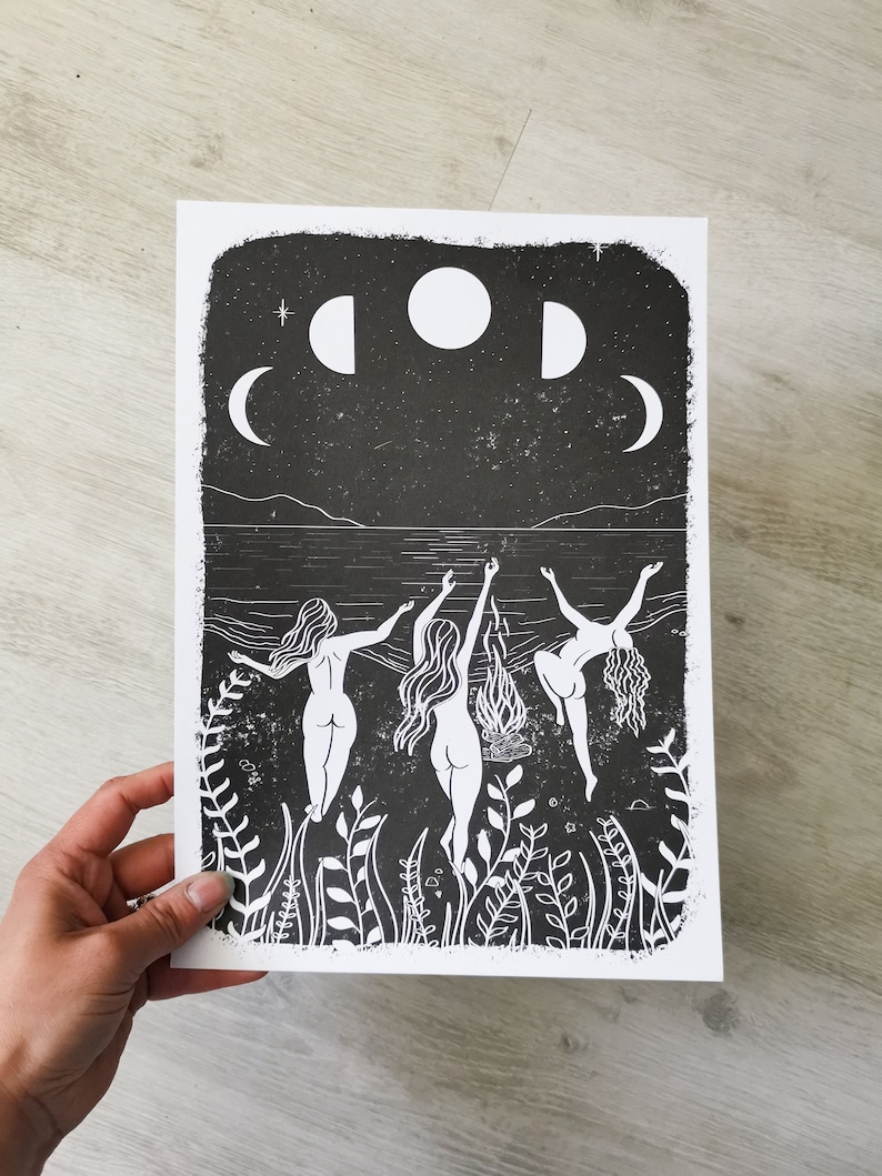 May include: Black and white linocut print of three women dancing in front of a bonfire under a night sky with three moons. The women are nude and have long hair. The print is framed by a border of plants.