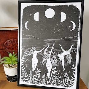 May include: Black and white print of three figures dancing in a circle around a fire under a crescent moon and stars. The figures are nude and have long hair. The print is framed in a black frame.