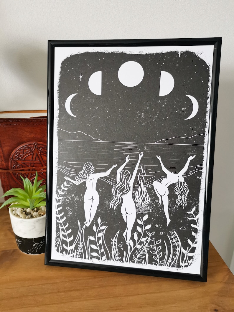 May include: Black and white print of three figures dancing in a circle around a fire under a crescent moon and stars. The figures are nude and have long hair. The print is framed in a black frame.