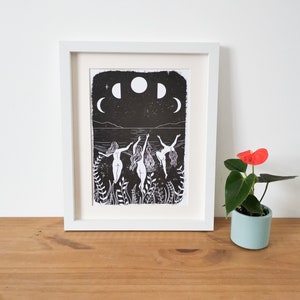 May include: Black and white print of three figures dancing in a body of water with a full moon and crescent moons in the sky. The print is framed in a white frame.