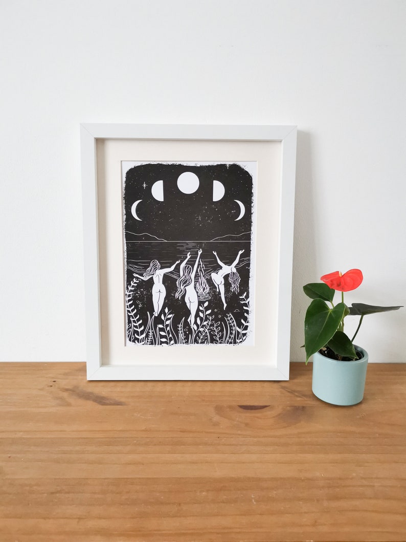 May include: Black and white print of three figures dancing in a body of water with a full moon and crescent moons in the sky. The print is framed in a white frame.