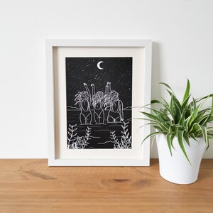 Op de afbeelding: Een wit ingelijst print met een zwarte achtergrond met een lijntekening van drie vrouwen die in het water staan met hun armen omhoog gericht naar de hemel. De maan en sterren zijn zichtbaar in de nachtelijke hemel.
