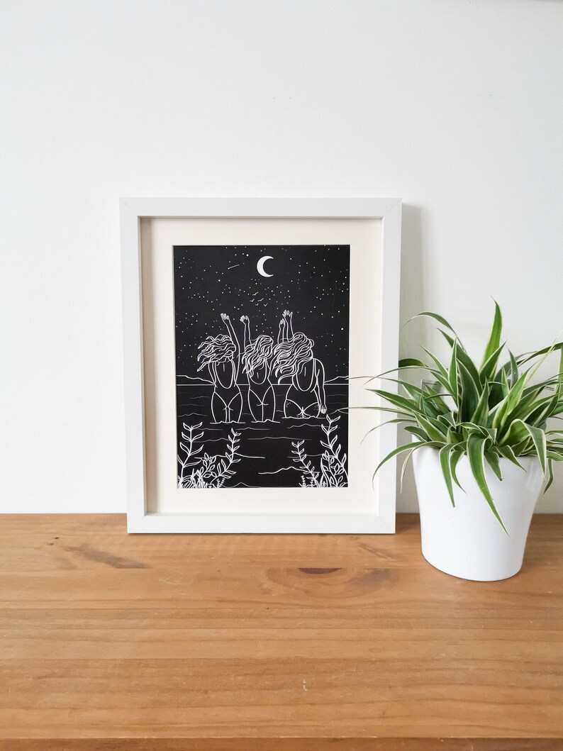 Op de afbeelding: Een wit ingelijst print met een zwarte achtergrond met een lijntekening van drie vrouwen die in het water staan met hun armen omhoog gericht naar de hemel. De maan en sterren zijn zichtbaar in de nachtelijke hemel.