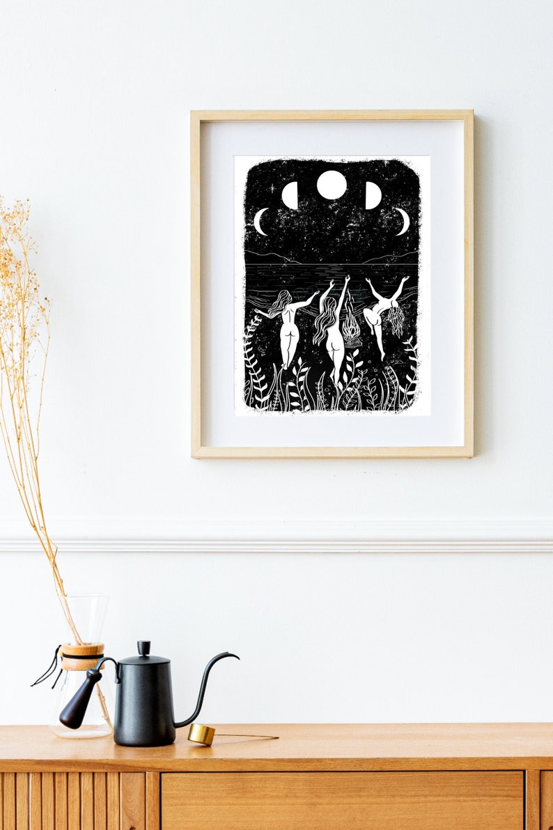 A4 art print on a white wall. The print is of three ladies dancing naked around a fire under the moon phases on the beach, surrounded by plants and leaves. Black & white print in lino style art.