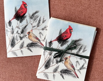 May include: Set of four greeting cards featuring watercolor illustrations of red cardinals perched on pine branches. The cards are tied together with a green ribbon.
