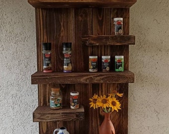 May include: A rustic wooden spice rack with multiple shelves. The rack is filled with various glass jars of spices and herbs. A small ceramic jug and a vase with sunflowers are also on the shelves.