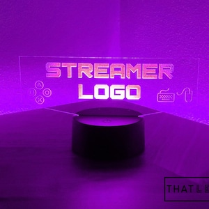 May include: A clear acrylic sign with the text "STREAMER LOGO" in a white font. The sign is illuminated with purple light and has icons for a controller, keyboard, and mouse.