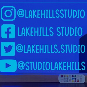 May include: A blue LED sign with a wooden base and a remote control. The sign displays social media handles for @lakehillsstudio, including Instagram, Facebook, Twitter, and YouTube.