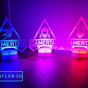 Puede incluir: Tres lámparas gamertag con diferentes diseños. Las lámparas tienen forma triangular con una base blanca y un panel de acrílico transparente. El texto gamertag está impreso en el panel. Las lámparas están iluminadas en diferentes colores: azul, rosa y rojo.