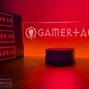 May include: A red LED sign with the text "GAMER+HAG" in white. The sign is on a black base and is lit up. The sign is in front of a stack of boxes with the text "THATLED.CO" on them.