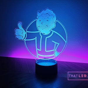Könnte beinhalten: Ein blaues und violettes LED-Nachtlicht mit einer Comicfigur, die einen Daumen nach oben zeigt. Die Figur ist weiß umrandet und befindet sich in einem Kreis. Das Licht steht auf einem schwarzen Sockel.