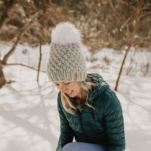 May include: A woman wearing a gray knit beanie with a white pom pom, a green puffer jacket, blue jeans, and brown suede boots. She is standing in the snow.