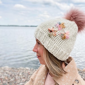 May include: A cream-coloured knitted hat with a large pink pom-pom and three small, colourful flower embellishments. The hat is made of thick yarn and has a textured appearance. The flowers are in shades of pink, yellow, and blue.