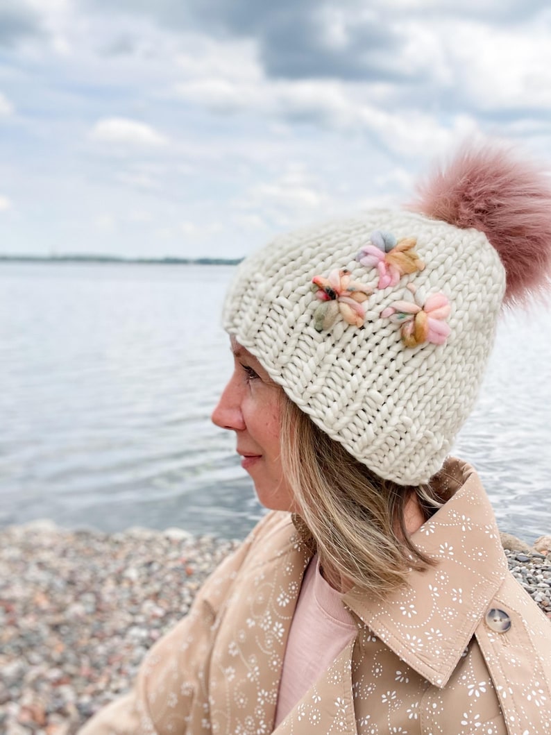 May include: A cream-coloured knitted hat with a large pink pom-pom and three small, colourful flower embellishments. The hat is made of thick yarn and has a textured appearance. The flowers are in shades of pink, yellow, and blue.