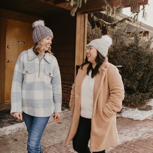 May include: Two women wearing winter clothing stand outside a building. One woman is wearing a blue and white plaid shirt, blue jeans, and a gray knit hat. The other woman is wearing a brown teddy bear coat, blue jeans, and a white knit hat. They are both smiling and looking at each other.